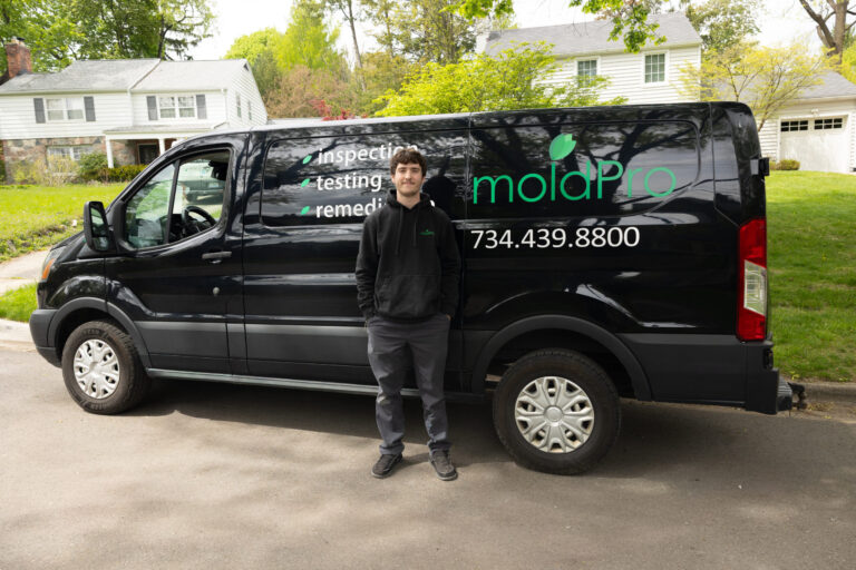 MoldPro technician standing beside the black MoldPro van, with the moldPro logo, phone number 734-439-8800, and inspection, testing, and remediation services listed on the side.